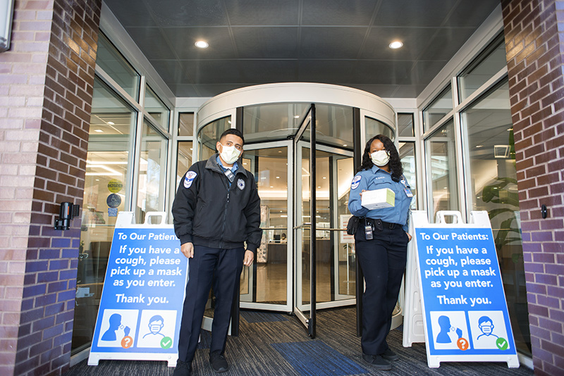 Two security officers hand out masks at the door at Englewood Health, next to signs that say "To our patients: if you have a cough, please pick up a mask as you enter. Thank you."