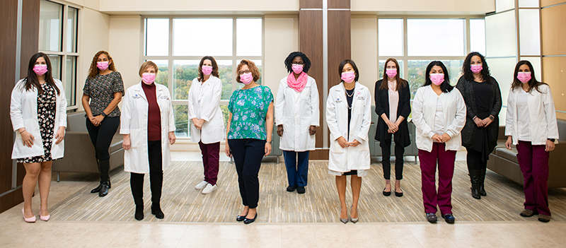 A group of Englewood Health team members in pink masks stand together in a room full of natural light.